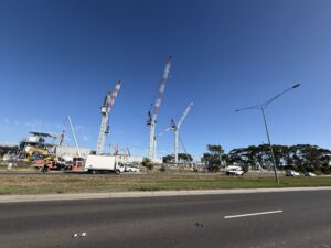 Construction site with multiple tall red-and-white cranes against a clear blue sky, trucks and equipment on the ground.